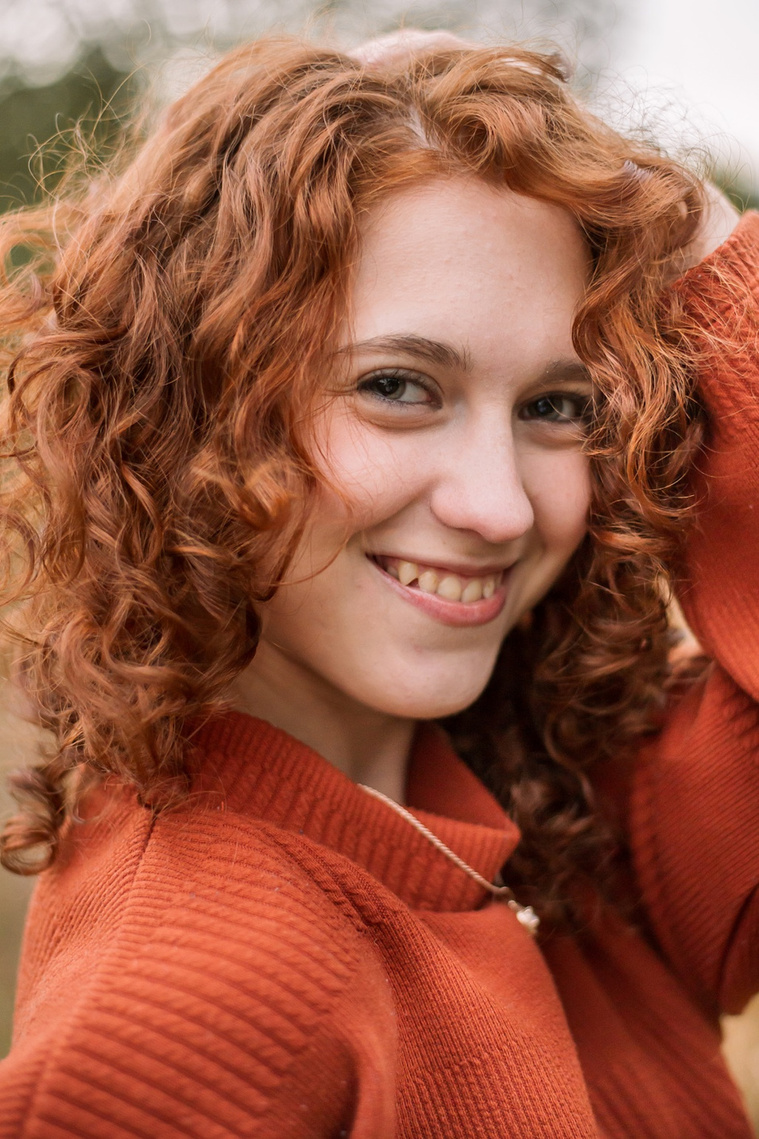 senior girl with red hair wearing an orange sweater close up photo with her hair framing her smiling face at Sutton Park in Norman, Oklahoma