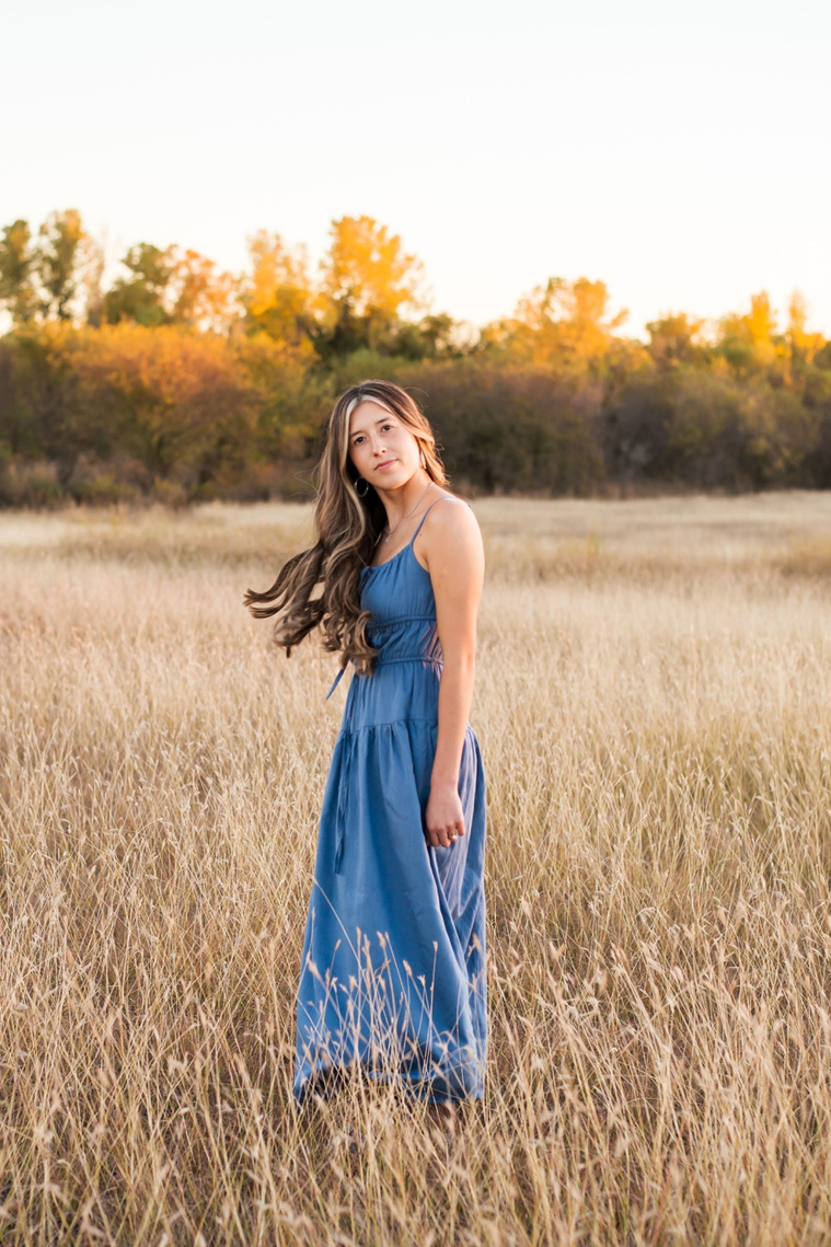 senior girl with long brown hair wearing a blue sundress stands to the side with her hair flowing behind her and smiling softly in a country field in the fall with trees behind her near Oklahoma City