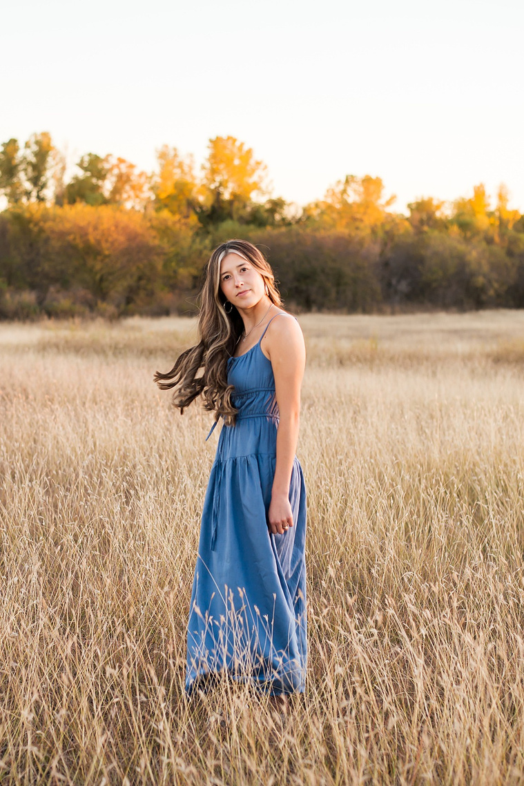 senior girl with long brown hair wearing a blue sundress stands to the side with her hair flowing behind her and smiling softly in a country field in the fall with trees behind her near Oklahoma City