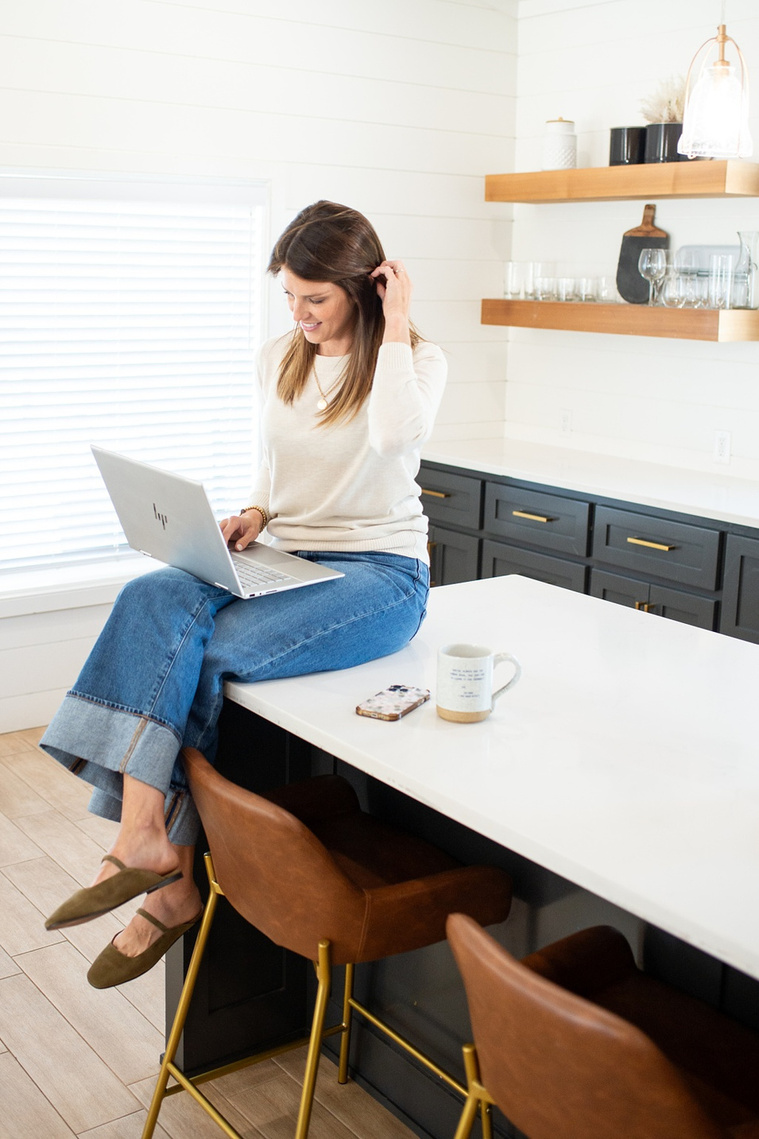Brand photo shoot of realtor in OKC. Natural image of her sitting on kitchen island with crossed legs working on her laptop with her phone and coffee cup by her