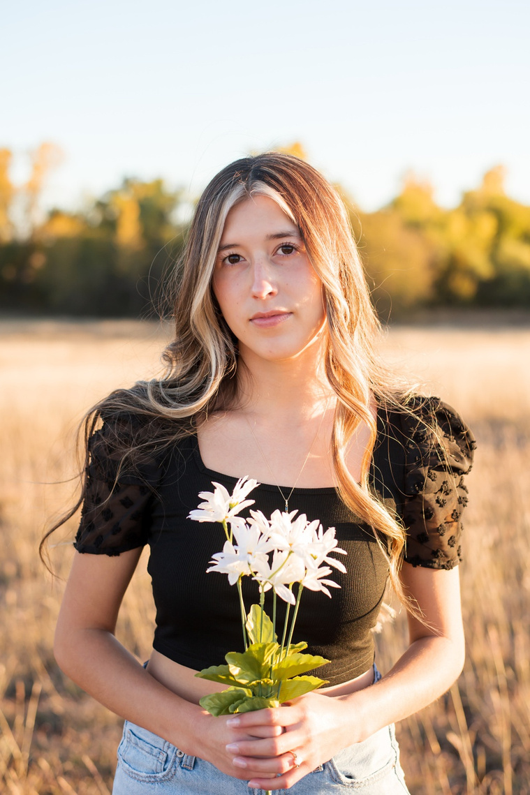 senior girl with long brown hair wearing a black top and jeans stands in tall grass holding a bouquet of white daisies with trees behind her, smiling softly in a country field in the fall near Oklahoma City