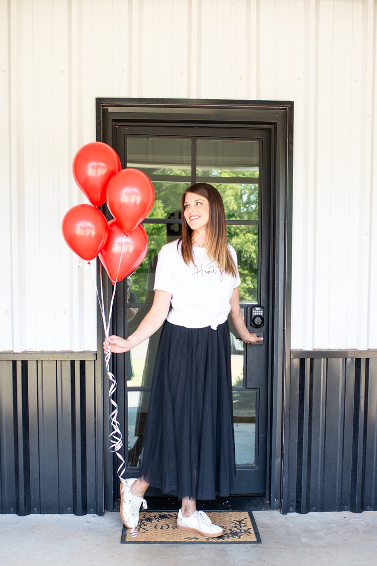 Brand photo shoot of realtor in OKC. She stands in front of entry door wearing black tulle skirt, t-shirt, tennis shoes holding a bunch of red balloons