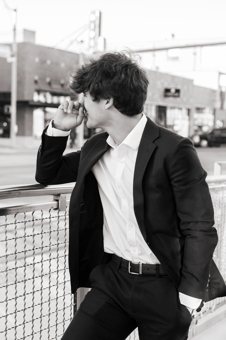 black and white portrait of a Hispanic high school senior boy wearing a dark blue suit and white shirt leaning on the street trolley fence and laughing away from the camera  in Automobile Alley in Oklahoma City