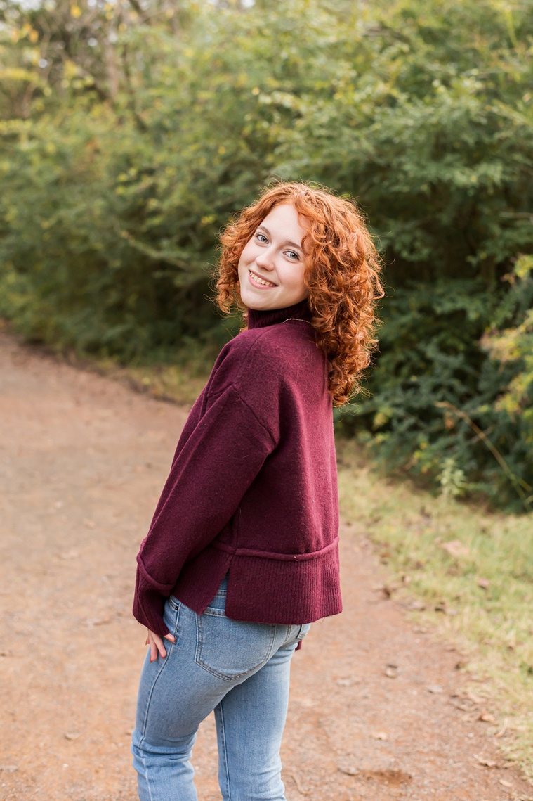 senior girl with red hair wearing a purple sweater and jeans looks cutely over her should smiling on a path with fall trees behind her at Sutton Park in Norman, Oklahoma