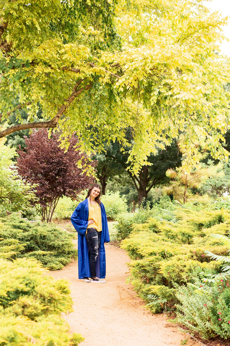 wide angle portrait shot of a high school senior girl with long brown hair wearing a yellow top and jeans and a blue graduation gown stands with hand on hip smiling with plants and tree leaves around her at Myriad Gardens in Oklahoma City