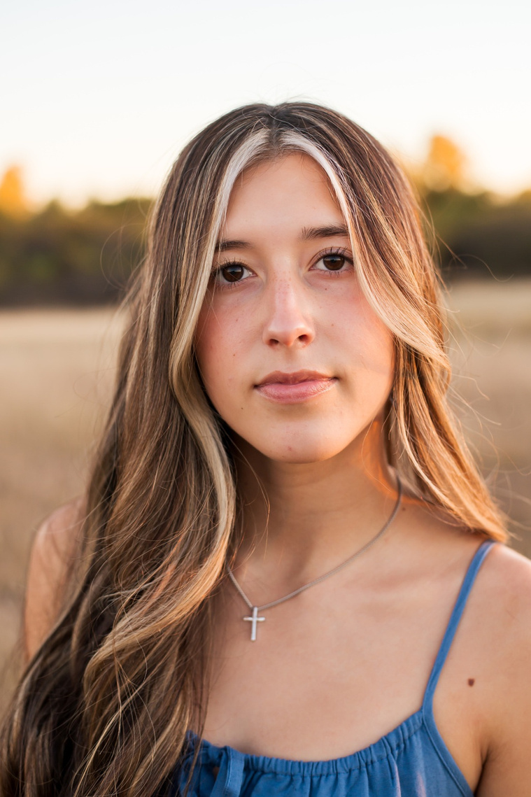 close up portrait of a senior girl with long brown hair wearing a blue sundress with a soft expression looking straight at camera standing in a country field in the fall near Oklahoma City