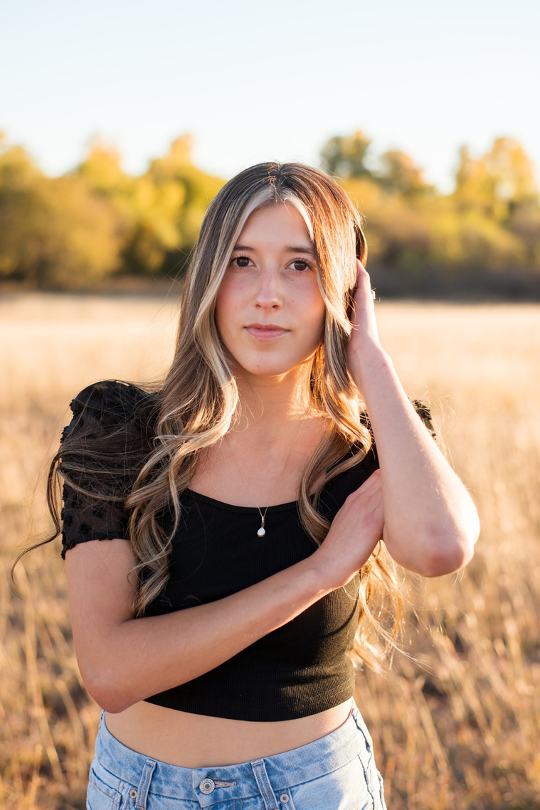 senior girl with long brown hair wearing a black top and jeans stands in tall grass with trees behind her, smiling softly with one hand in her hair in a country field in the fall near Oklahoma City