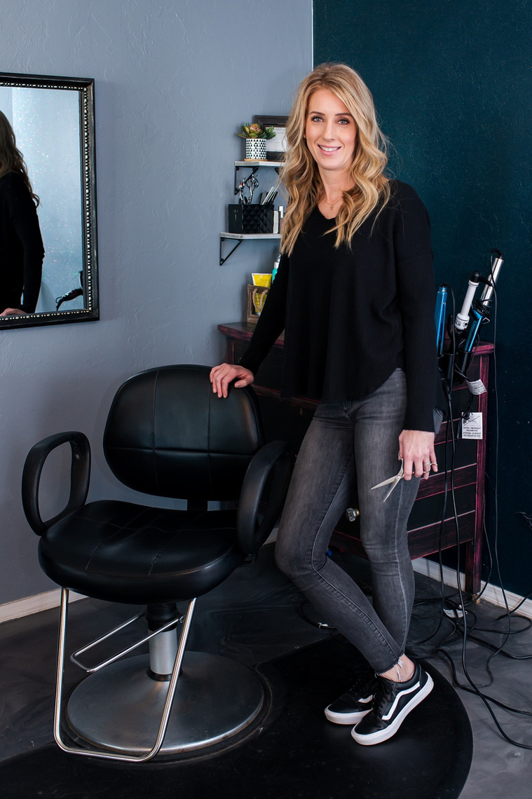 vertical full length portrait of a blond hair stylist in a black shirt and jeans standing by her chair smiling and holding her scissors at a branding session at Vibe Beauty Bar in Tuttle, Oklahoma