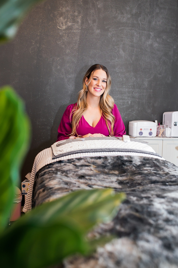 an esthetician in a pink shirt sits at the head of her client bed smiling with green plants in foreground her facial supplies around her at a branding session at Vibe Beauty Bar in Tuttle, Oklahoma