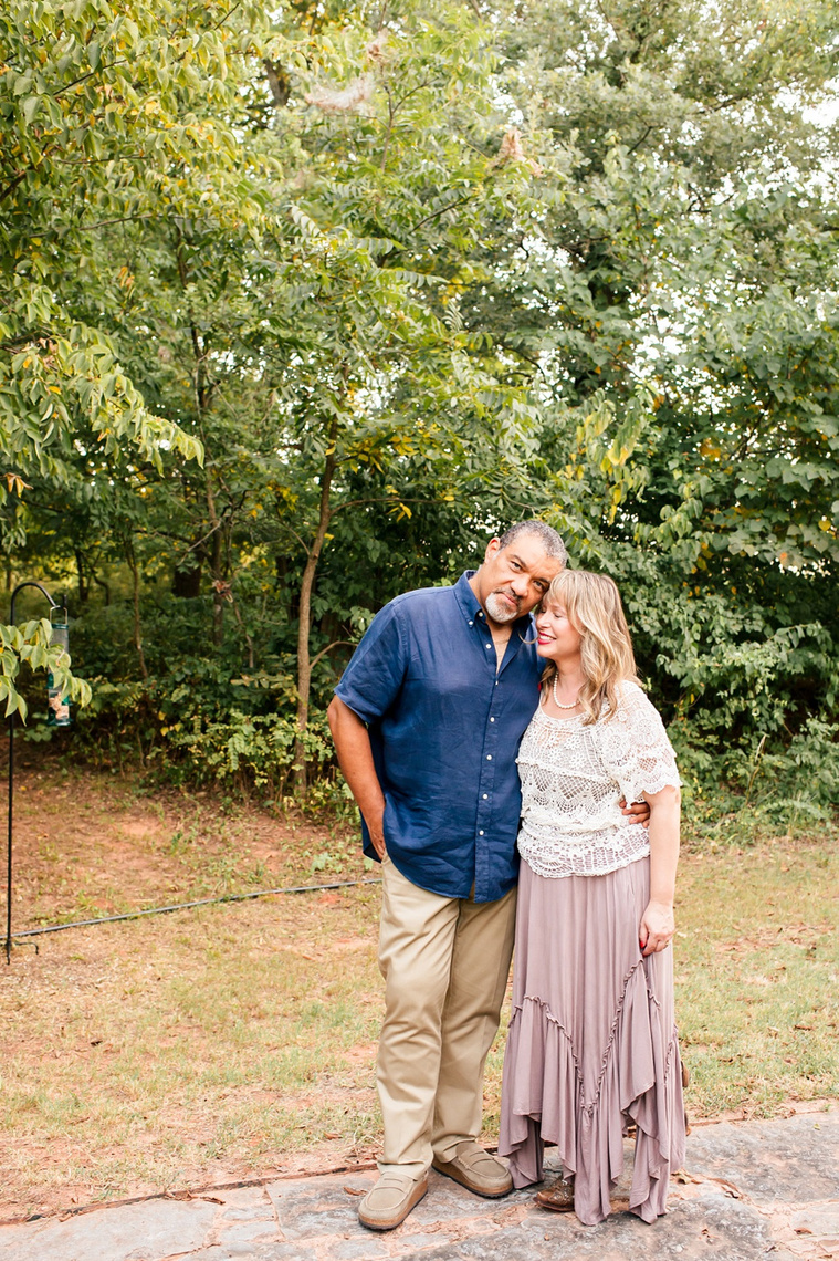 a middle aged husband and wife stand together closely leaning into each other with trees behind them at their home in Norman, Oklahoma