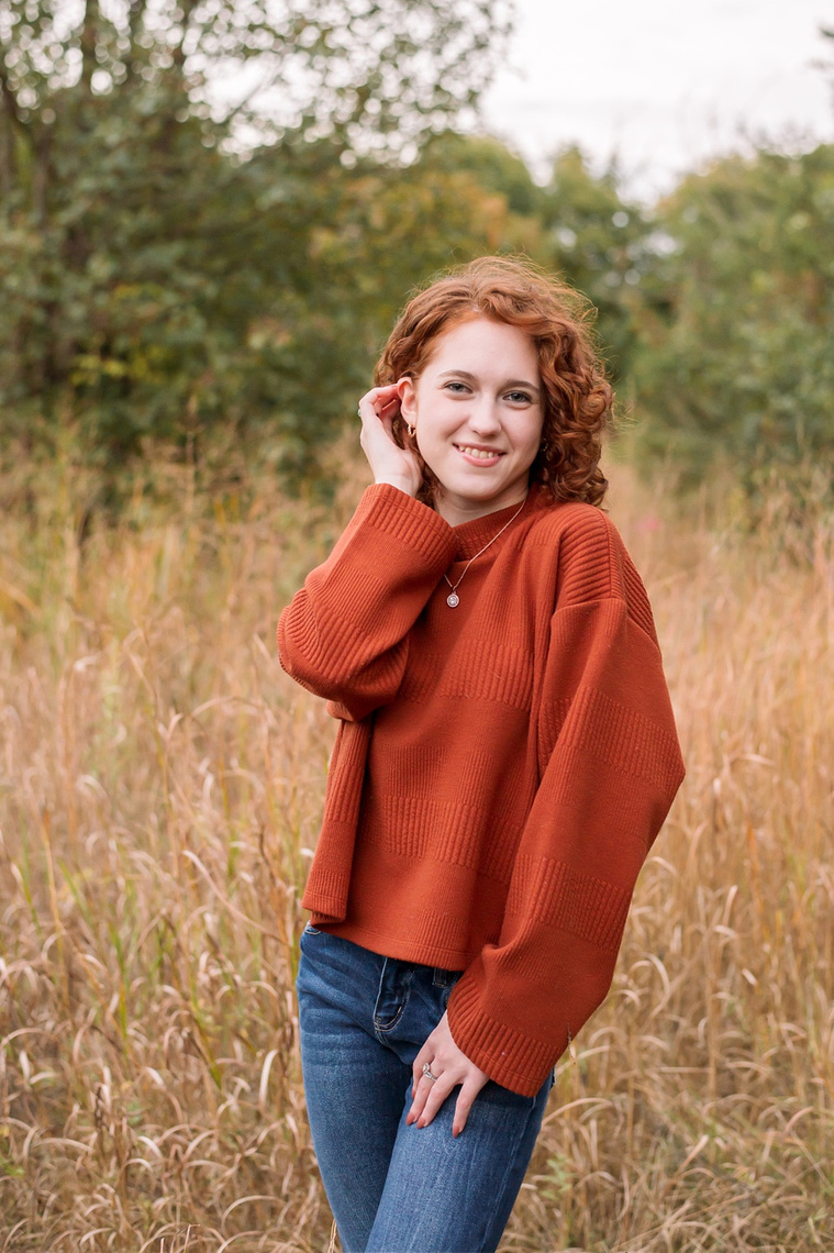 senior girl with red hair wearing an orange sweater and jeans poses cutely and smiles standing in a field with gold grass and fall trees behind her at Sutton Park in Norman, Oklahoma
