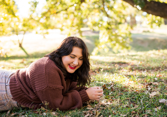 High school senior girl lays on her stomach propped on her elbows, laughing in the grass with trees and leaves around her at a fall senior photo session at Will Rogers Gardens in Oklahoma City