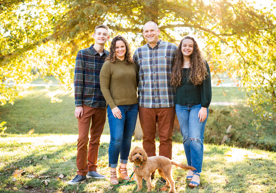 A family of four stand with their medium size dog in a park with glowy-light trees behind them for a family photo by Cheryl Jackson Photography at Will Rogers Gardens in Oklahoma City