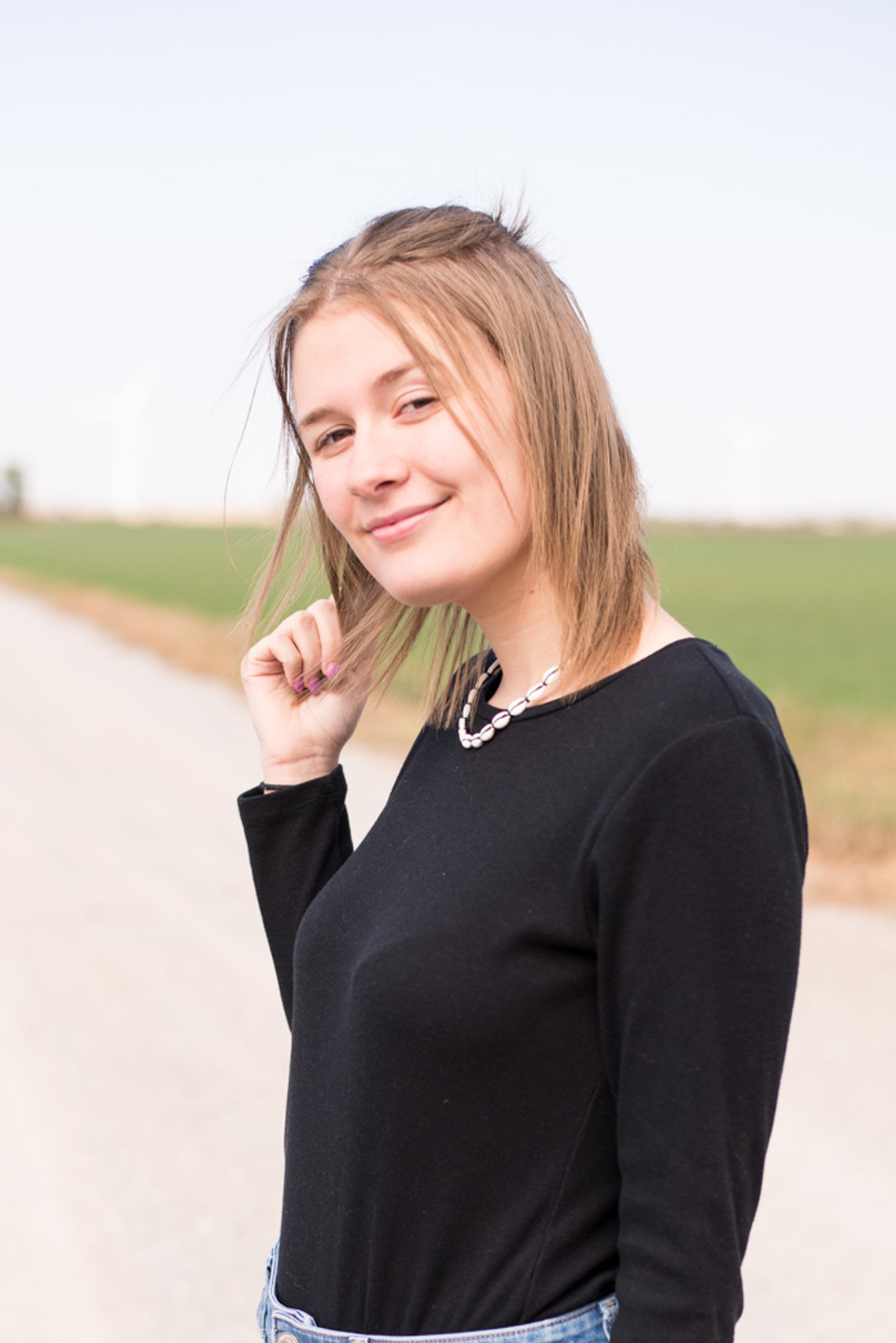 Portrait of a teenage girl in a black t-shirt and jeans standing sideways to camera with one hand in her hair and a soft smile on a dirt road