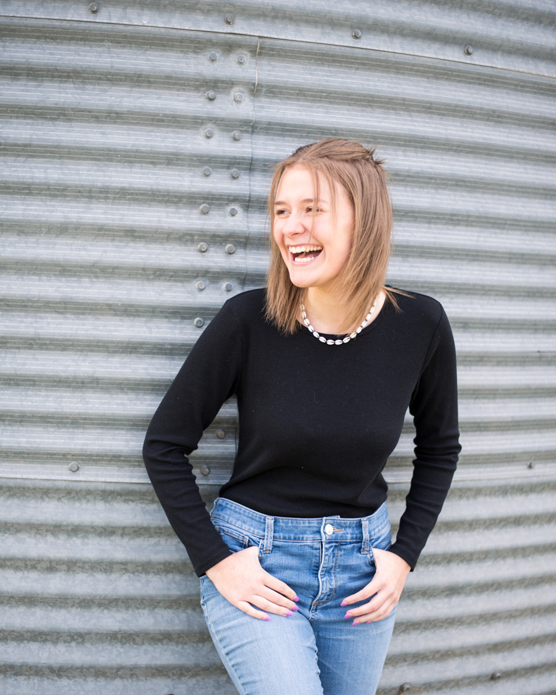 Portrait of a teenage girl in a black t-shirt and jeans in front of grain bin, looking off to the side and laughing