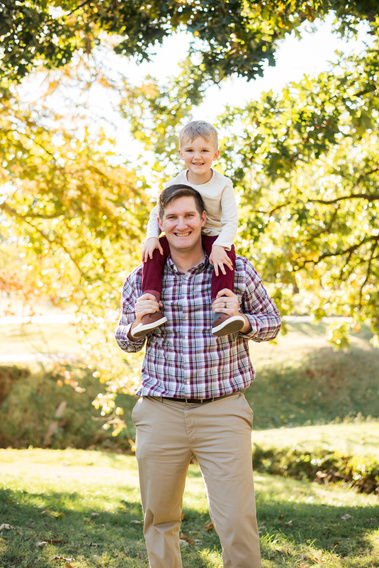 a dad smiles while his young son sits around his neck and smiles with fall trees and sunlight behind them at Will Rogers Gardens in Oklahoma City
