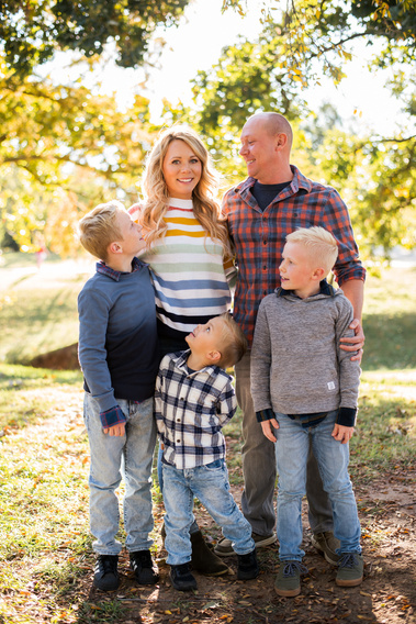 a mom and dad with their 3 young sons stand together hugging and looking at one another with smiles in front of fall trees at Will Rogers Gardens in Oklahoma City