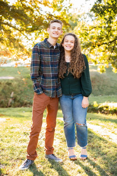 a teen brother and sister wearing earthy toned clothing stand and smile together with fall trees behind them at Will Rogers Gardens in Oklahoma City