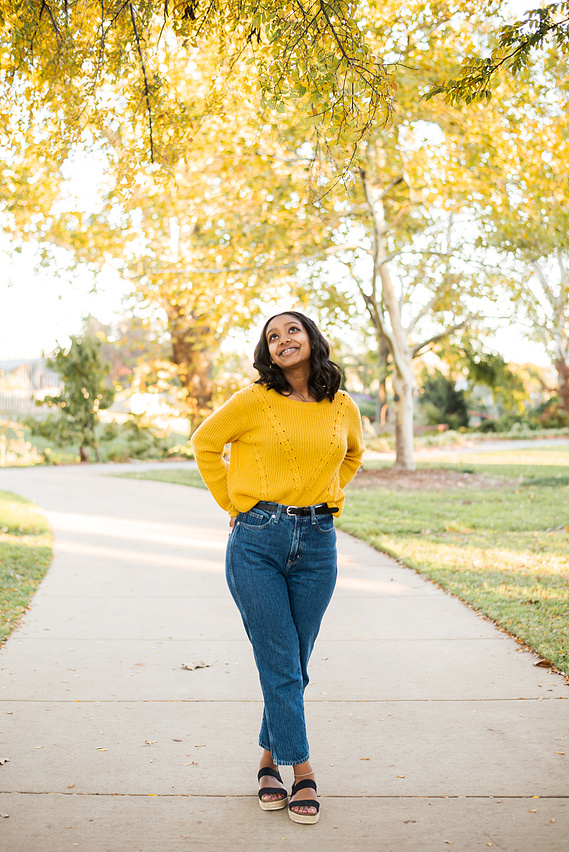 senior girl wearing a golden yellow sweater and jeans stands cutely on a sidewalk looking up at golden fall leaves above her at Will Rogers Gardens in OKC