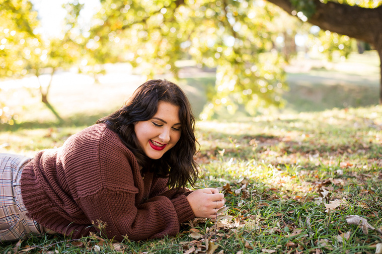 High school senior girl lays on her stomach, laughing, propped on her elbows in the grass with trees behind her at Will Rogers Gardens in Oklahoma City for her fall senior photo session