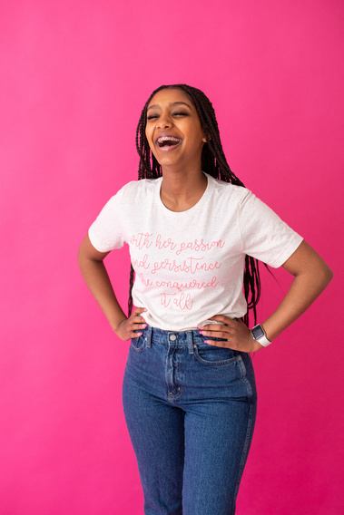 a teen girl with long braids stands with her hands on her hips and laughs in front of a pink seamless backdrop