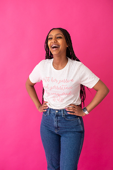 a teen girl with long braids stands with her hands on her hips and laughs in front of a pink seamless backdrop