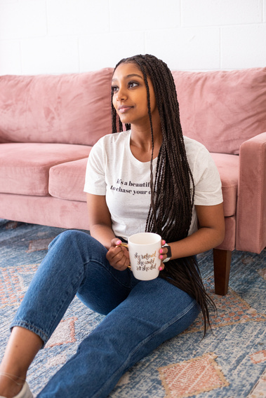 a teen girl with long braids sits  on the floor on a rug in front of a pink couch holding a coffee mug