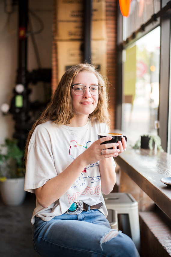 senior portrait of a girl with glasses and long blond hair wearing a vintage tee and ripped jeans holding a cup of coffee along the outer windows of a coffee shop in midtown Oklahoma City