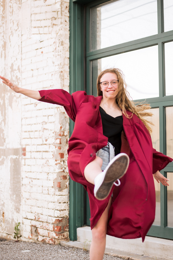 senior girl wearing her graduation gown kicks forward playfully with her arms outstretched and smiles
