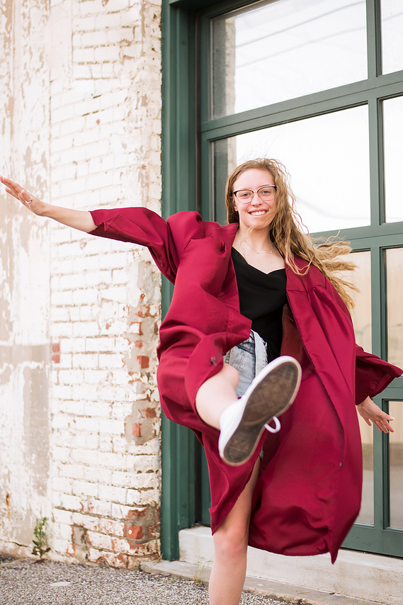 senior girl wearing her graduation gown kicks forward playfully with her arms outstretched and smiles