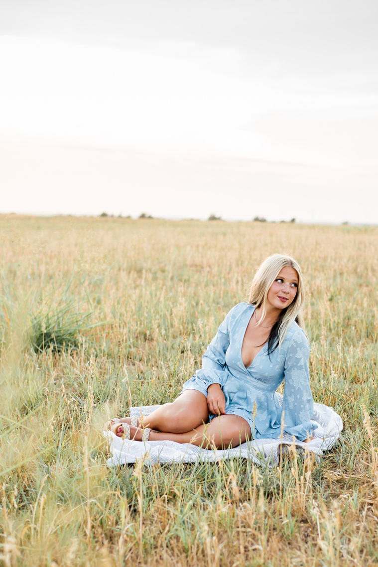 High school senior girl sits on a blanket in the middle of a field and looks off to the side with a knowing smile in a blue dress in a field in central Oklahoma for her senior photo session