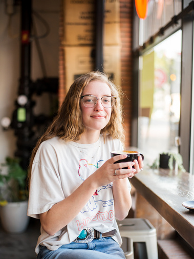 high school senior girl sits in a coffee shop holding a coffee mug and smiles at a senior portrait session