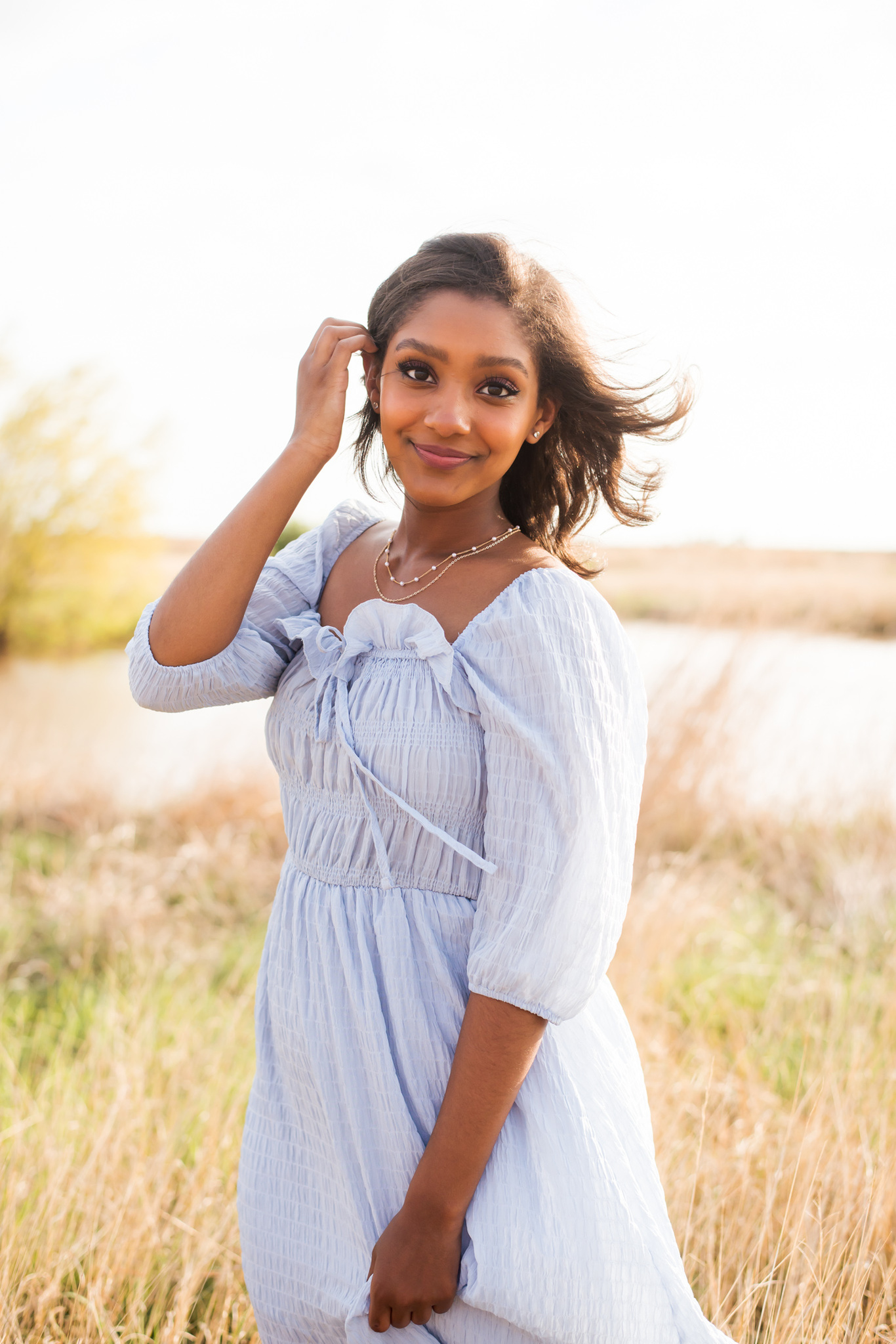 black high school senior girl wearing a blue dress stands in a field with a pond behind her tucking her hair behind her ear and smiling softly near Tuttle Oklahoma