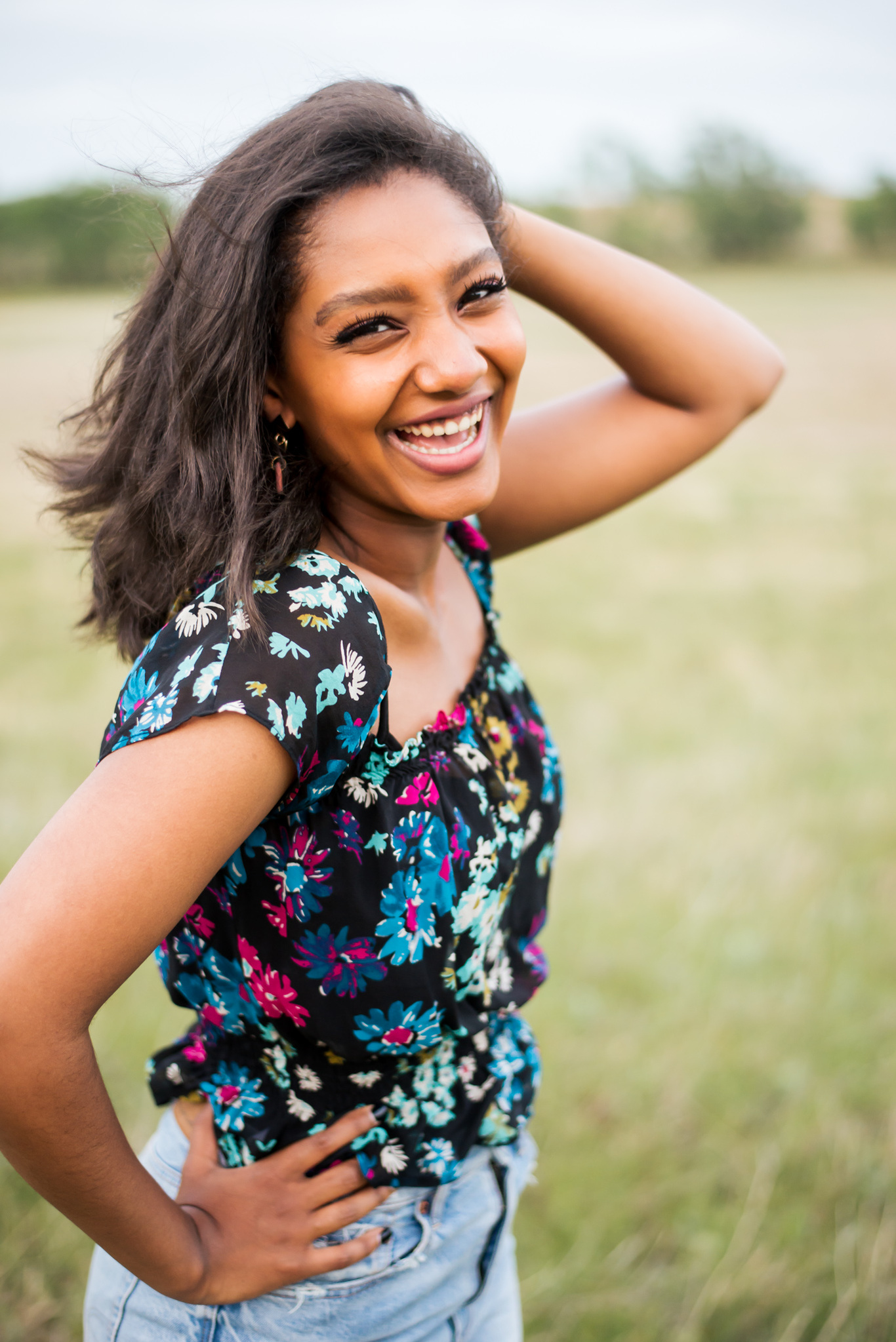 portrait of a african american high school senior girl with one hand on her hip and the other in her hair laughing at the camera wearing jeans and a floral top while she stands in a field in central Oklahoma