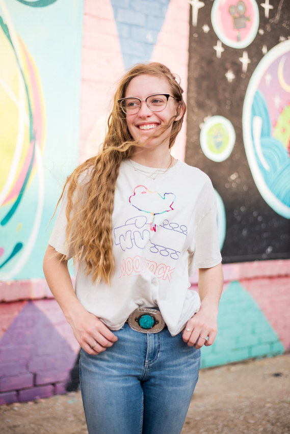 Blond high school senior girl with glasses smiles off camera with her thumbs in her belt loops standing in front of a graffiti wall at Factory Obscura in midtown Oklahoma City for her senior photo session