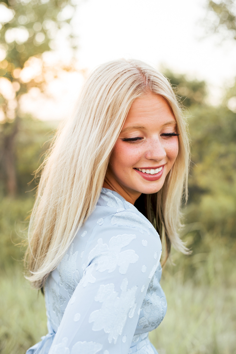 High school senior girl smiling over her shoulder and looking down slightly in a blue dress in a field in central Oklahoma for her senior photos