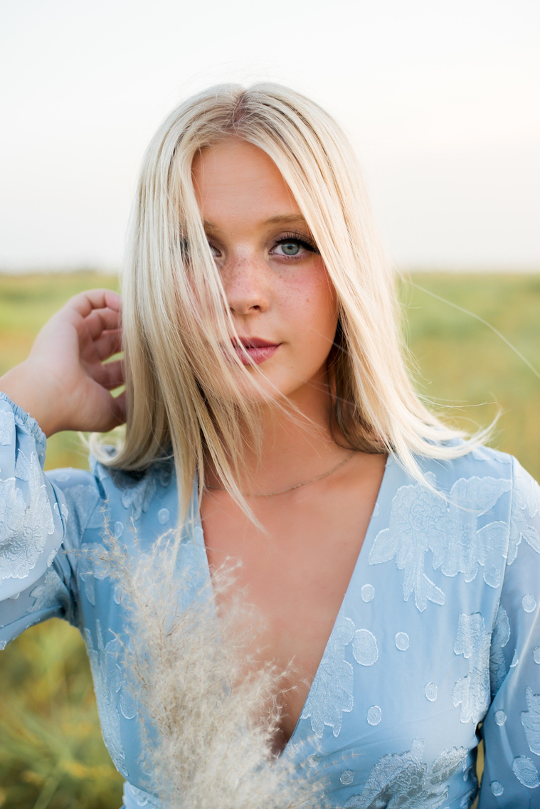 High school senior girl in a blue dress is looking at camera with a soft smile while the wind blows her blond hair over half her face in a field in central Oklahoma for her senior photos