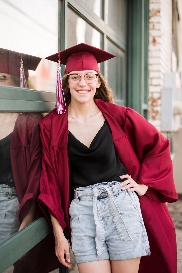 high school senior girl wearing a red cap and gown leans against a wall of windows framed in green and smiles for her senior photos in Oklahoma City