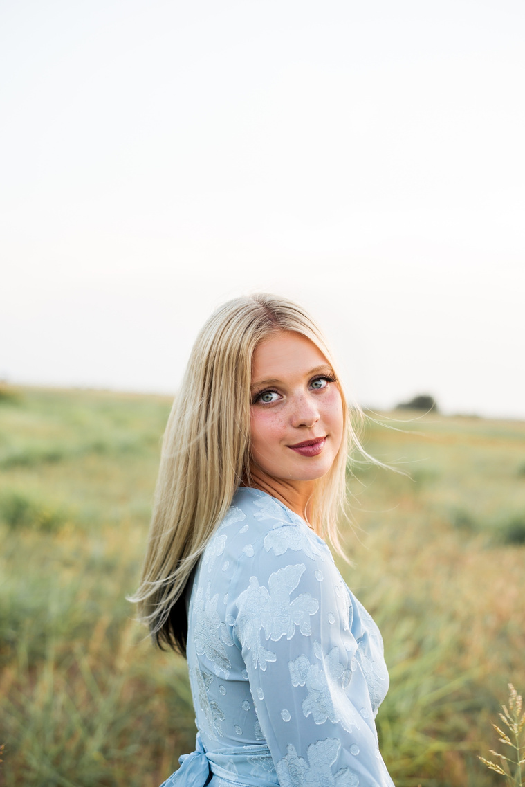 High school senior girl looks over her shoulder and softly smiles in a blue dress in a field in central Oklahoma for her senior photos