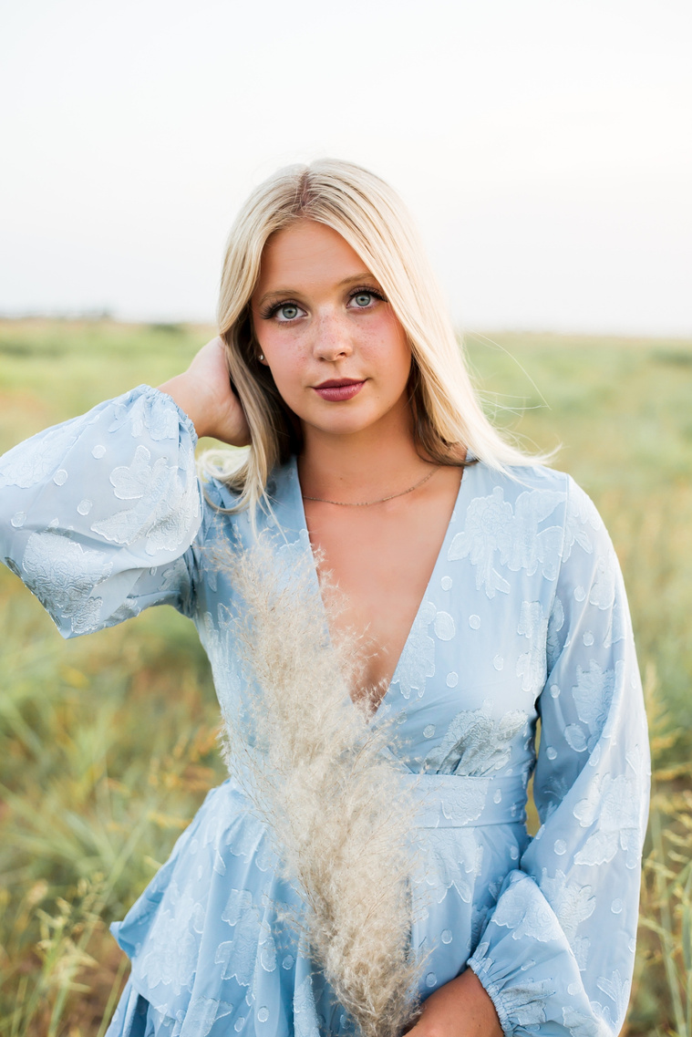 High school senior girl stands with one hand in her hair and the other holding a frond of pampas grass in a blue dress in a field in central Oklahoma for her senior photos