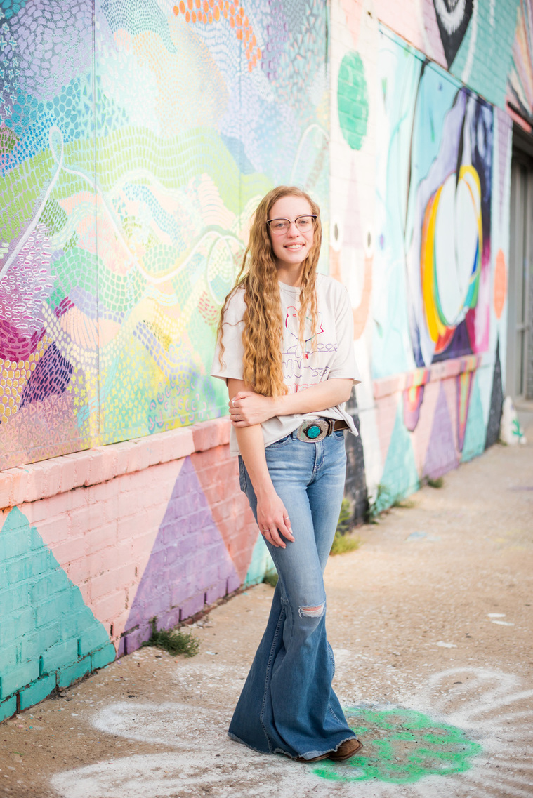 senior girl with long hair wearing 70s style clothes stands casually in front of a colorful mural wall in midtown OKC
