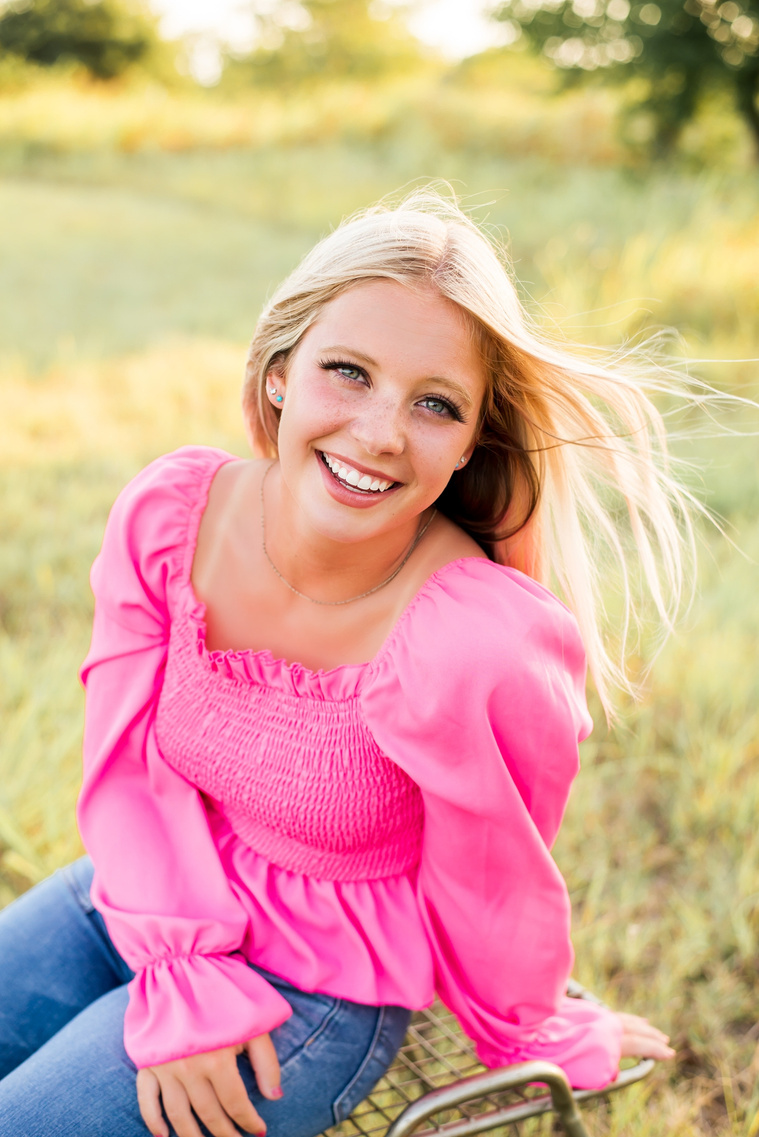 Joyful image of a high school senior girl sitting in a field in central Oklahoma and smiling happily in a pink top at her senior photo shoot