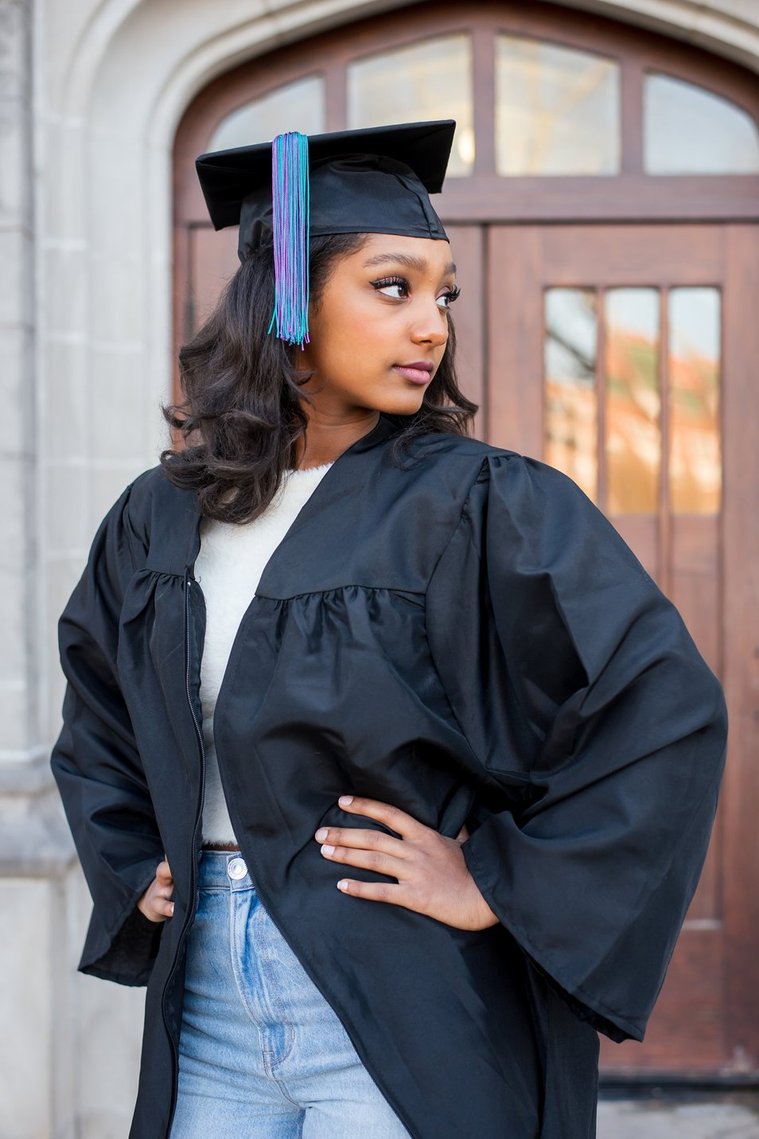 portrait of an african american high school senior girl wearing a black graduation cap and gown with tassel, looking off to the side seriously with her hands on her hips in front of doors at a building on the OU campus in Norman Oklahoma