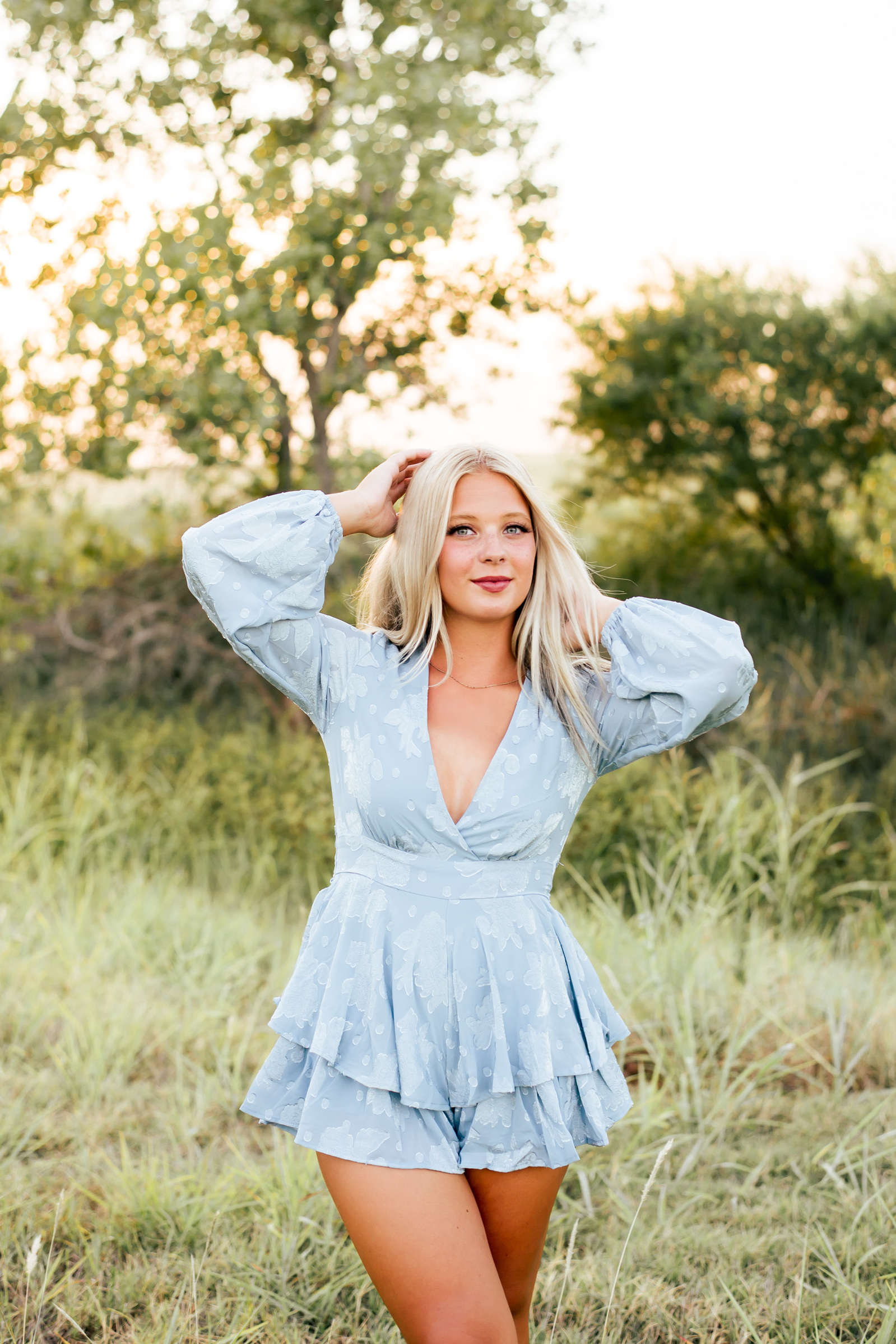 high school senior girl with blond hair wearing a blue short jumper stands in a model pose with legs crossed and arms up and in her hair in a field in Oklahoma