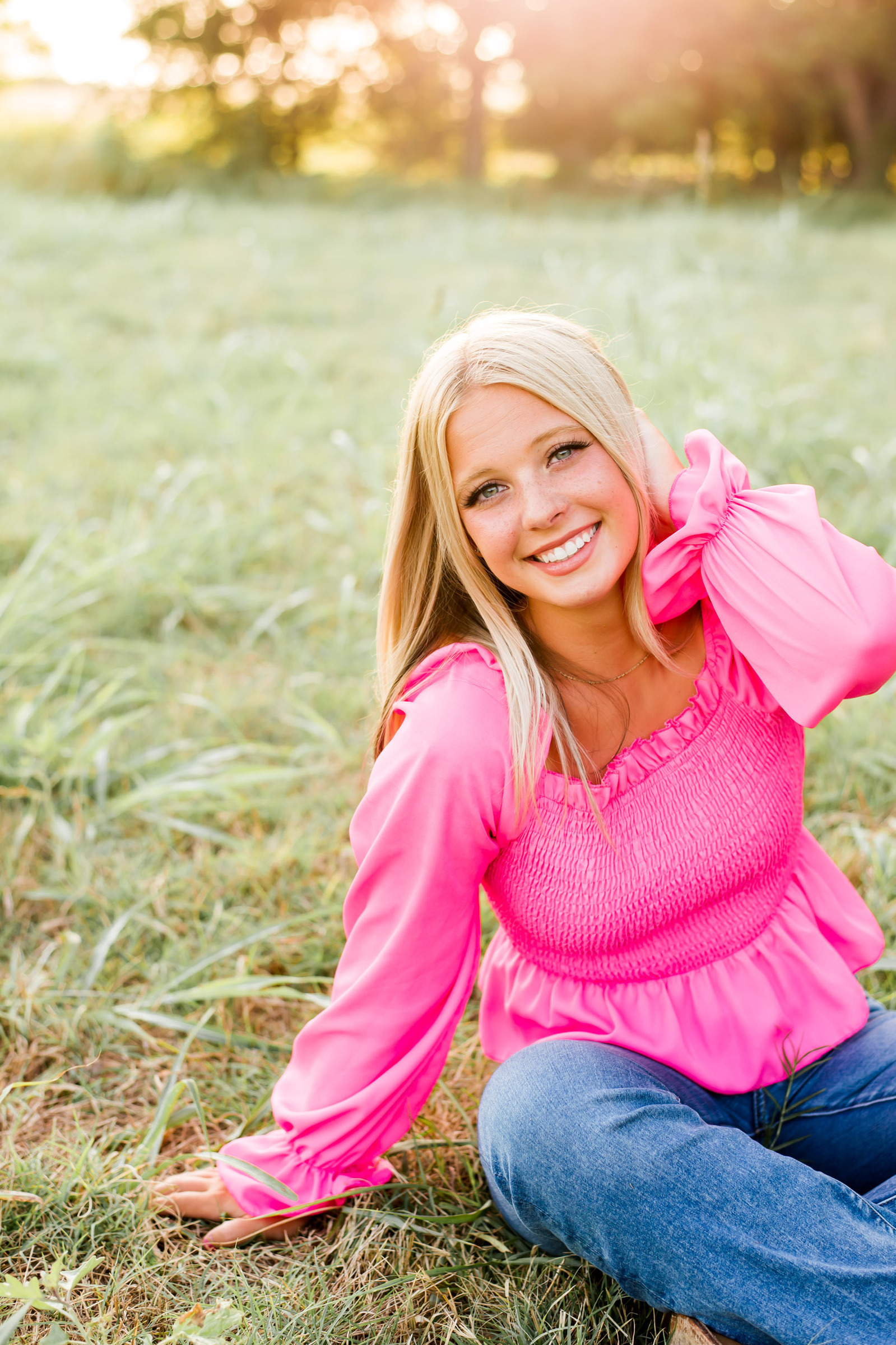 blond high school senior girl wearing a bright pink ruffle top and blue jeans sits and smiles in a field with golden light behind her