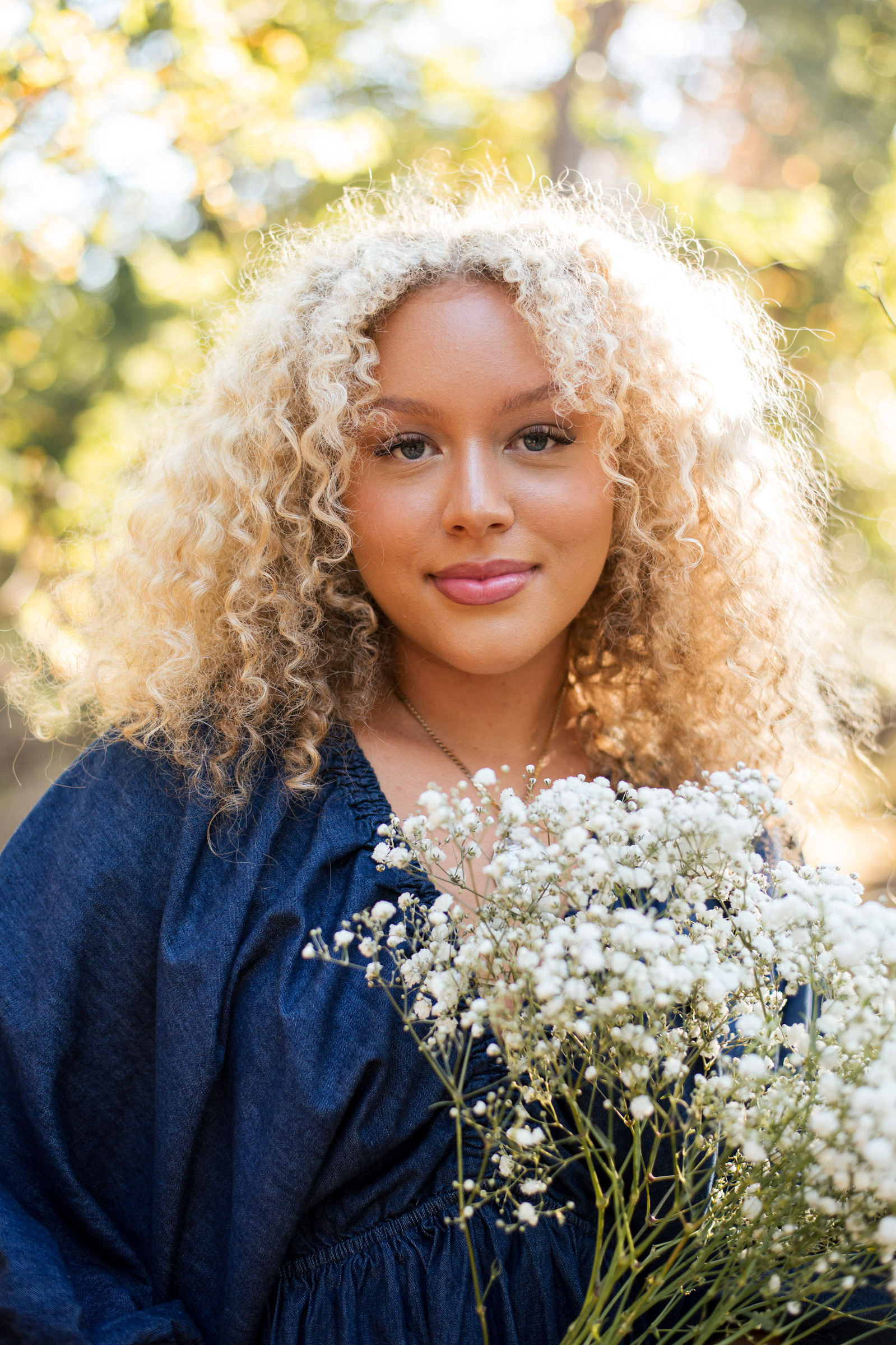 senior girl with curly blond hair holding a bouquet of flowers with sunlit leaves behind her smiles softly for senior pictures at Martin Nature Park in Oklahoma City, taken by OKC senior photographer