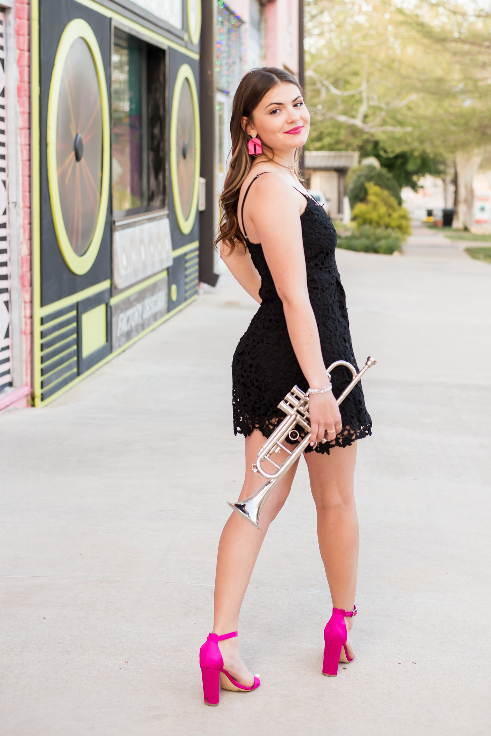 senior girl wearing a short black dress and bright pink heels looks over her shoulder holding her trumpet on a sidewalk in front of Factory Obscura in OKC