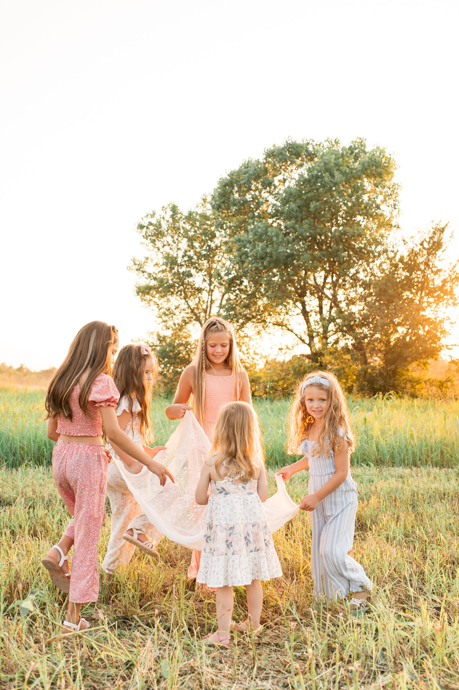 five young girls hold a piece of fabric and dance in a circle at sunset in a country field with a few trees behind them