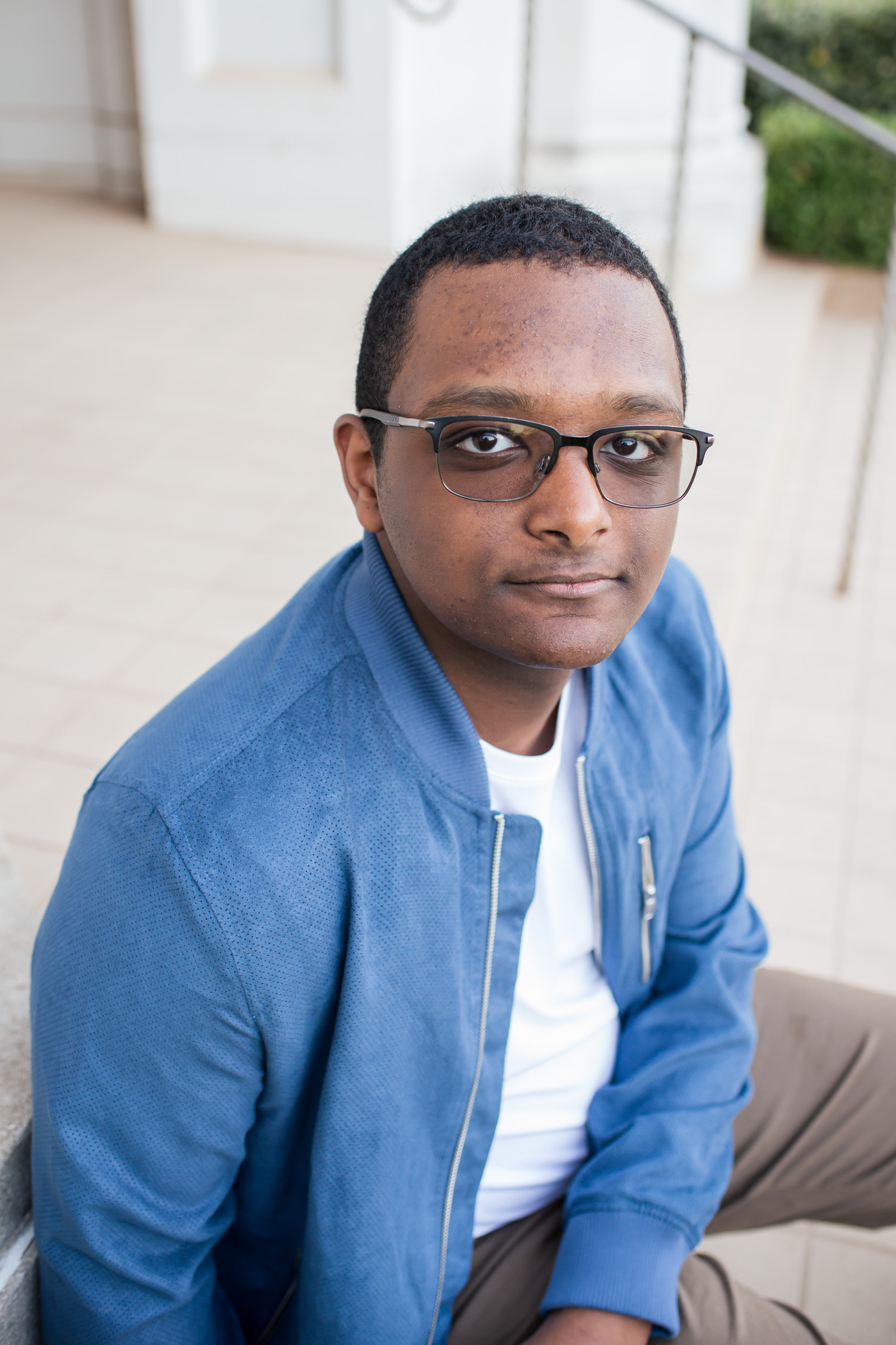 portrait of a black high school senior boy wearing a bright blue jacket with white shirt underneath, sitting on steps at OU campus building in Norman, Oklahoma