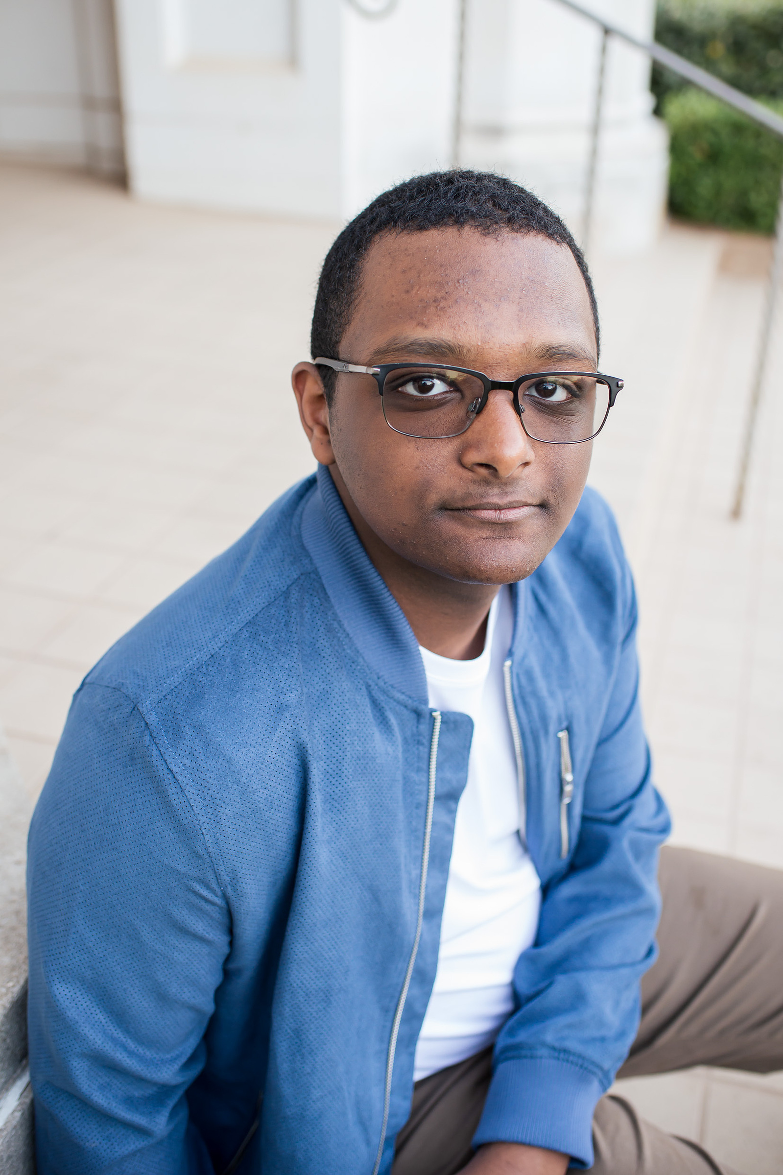 portrait of a black high school senior boy wearing a bright blue jacket with white shirt underneath, sitting on steps at OU campus building in Norman, Oklahoma